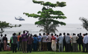 Dozens of bystanders look on as they watch rescuers in a helicopter search the site of a capsized cruise boat on Lake Victoria near Mutima village, south of Kampala, Uganda, on a grey, cloudy day.