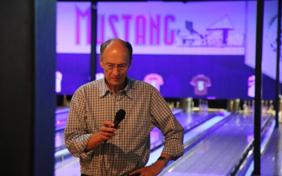 Roger McMacken stands holding a microphone, with the Mustang bowling lanes behind