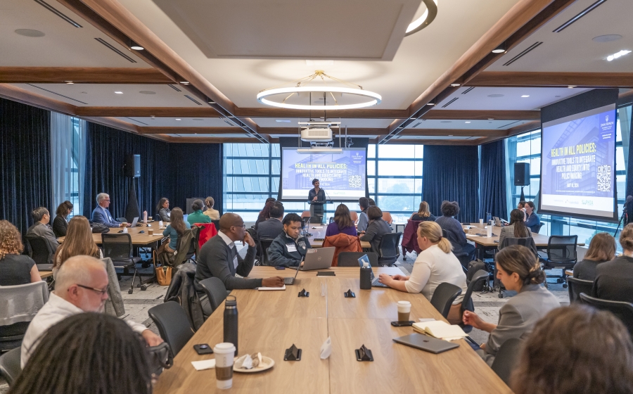 A diverse group of people seated at several long wooden tables, some with laptops or coffee cups by their arms. Most are facing towards the front of the wide room towards a woman, Keshia Pollack Porter, who is standing in front of a projector screen.