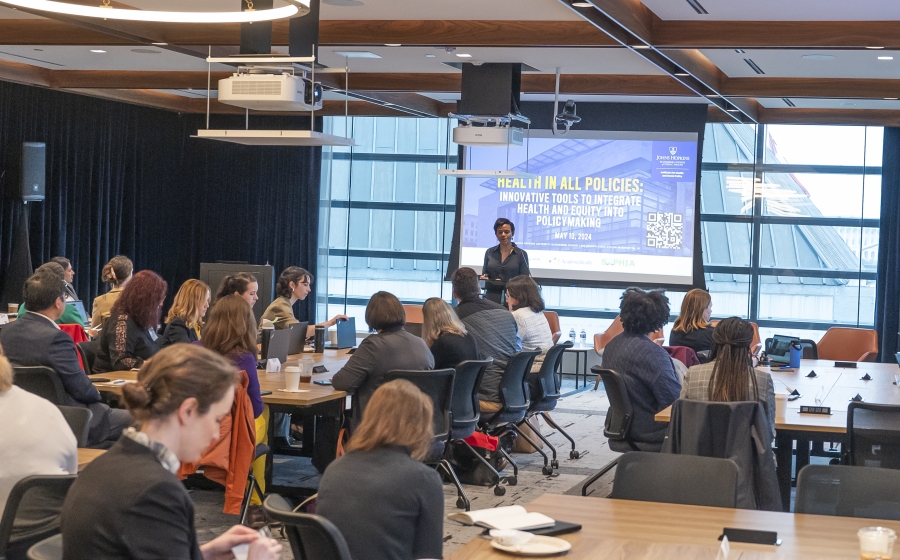 A diverse group of people seated at several long wooden tables, some with laptops or coffee cups by their arms. Most are facing towards the front of the wide room towards a woman, Keshia Pollack Porter, who is standing in front of a projector screen.