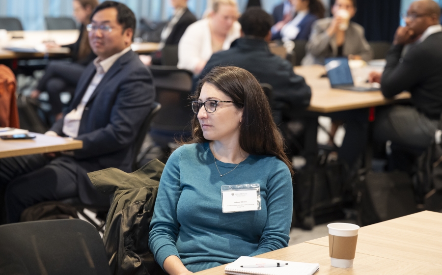 A woman in a teal long-sleeve shirt is seated at a wooden table with a coffee cup near her. Her attention is towards front of the room. In the background, multiple people at other tables are facing the same direction.