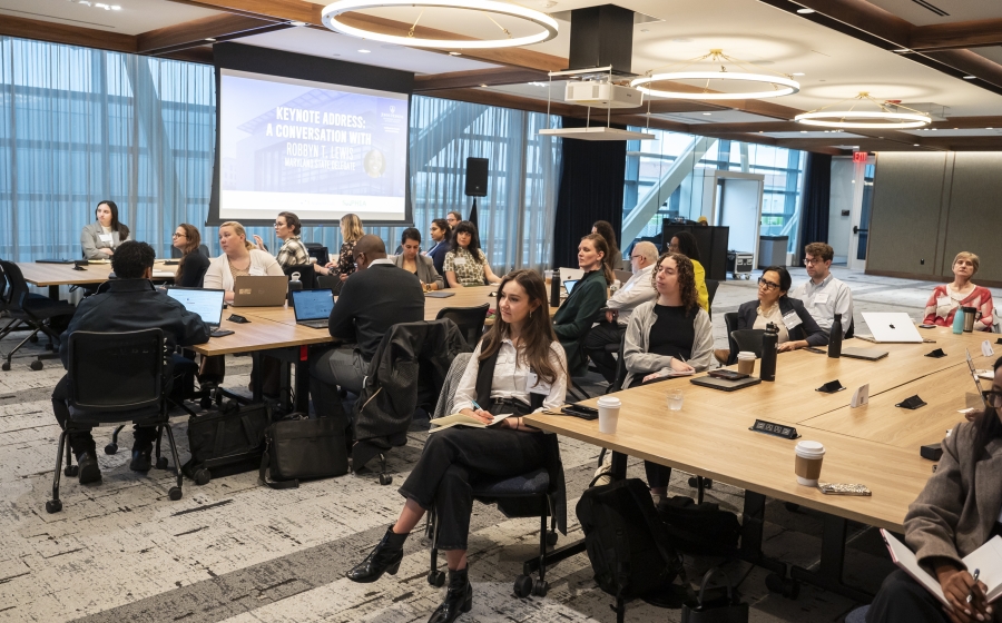 A racial and gender diverse group of people seated at several long wooden tables, some with laptops or coffee cups by their arms. Most are facing towards the front of the wide room.