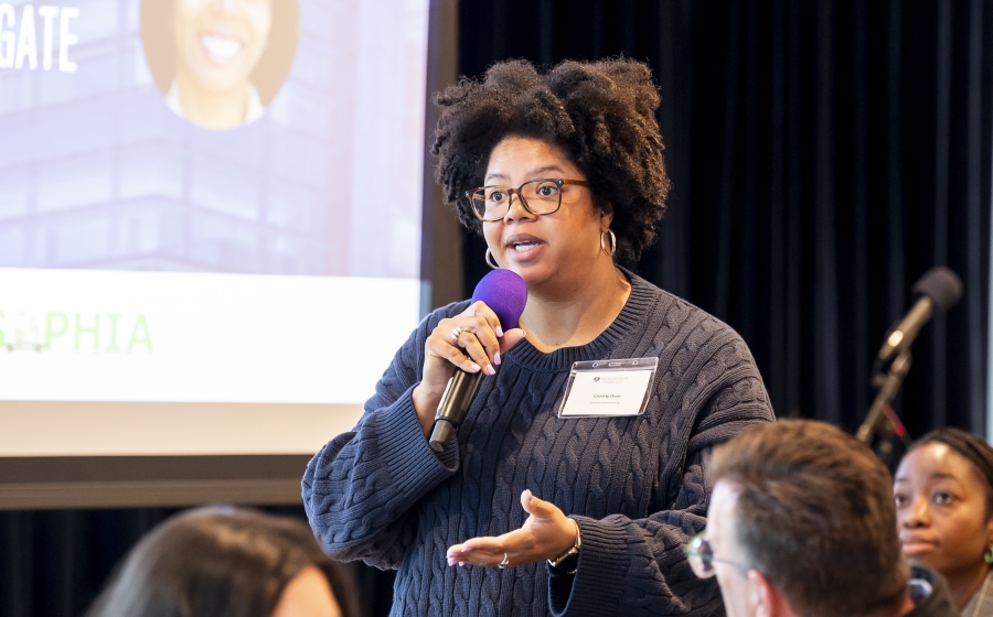 A woman wearing a black sweater stands while holding a hand-held microphone. She's speaking towards the front of the room, which is out of frame. A corner of a projector screen is visible in the background.