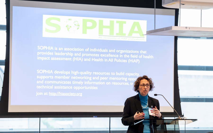 A woman stands behind a podium and in front of a projector screen. The title on the screen reads SOPHIA. The first sentence below the title reads SOPHIA is an association of individuals and organizations that provides leadership and promotes excellence in the field of health impact assessment (HIA) and health in all policies (HiAP).