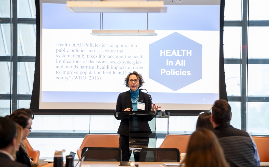 A woman stands behind a podium and in front of a projector screen. The title on the screen reads Health in All Policies. The first sentence next to the title reads Health in All Policies is "an approach to public policies across sectors that systematically takes into account the health implications of decisions, seeks synergies and avoids harmful health impacts in order to improve population health and health equity" (WHO, 2013).