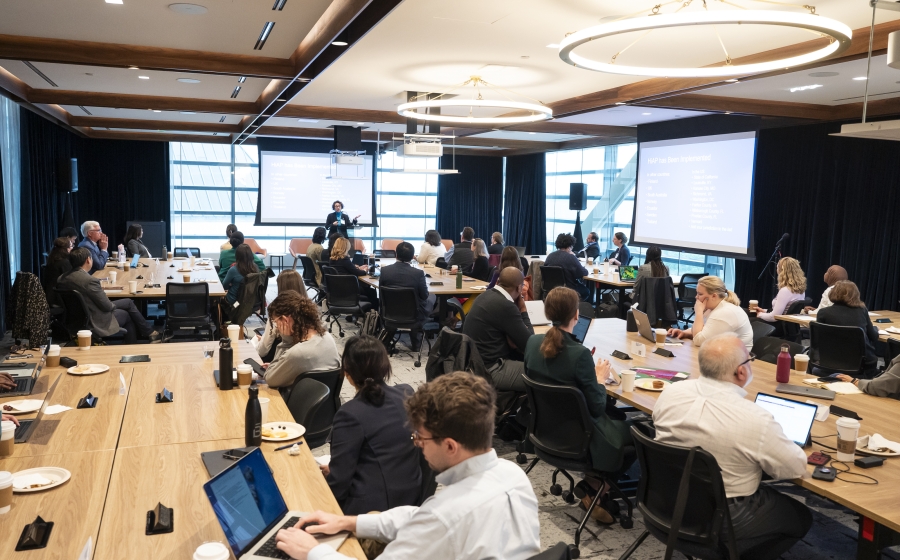 A racial and gender diverse group of people seated at several long wooden tables, some with laptops or coffee cups by their arms. Most are facing towards the front of the wide room.