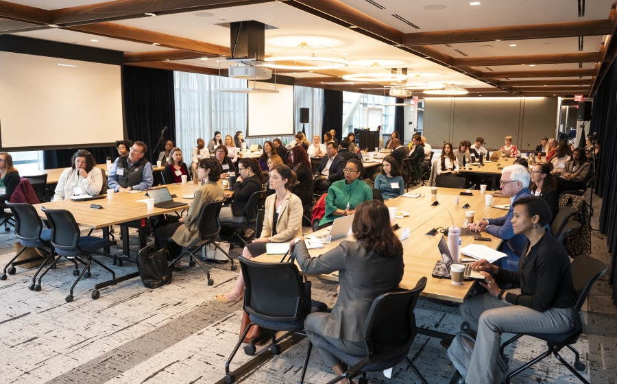 A racial and gender diverse group of people seated at several long wooden tables, some with laptops or coffee cups by their arms. Most are facing towards the front of the wide room.