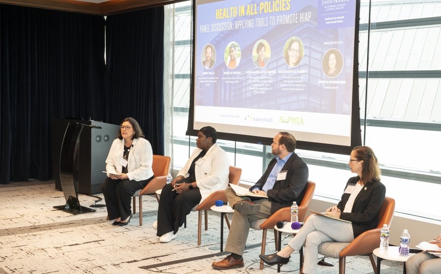 Four women and one man are seated in a row at the front of the room with a projector screen behind them. At the far right, the fourth woman is partially out of frame. The screen is entitled Health in All Policies Panel Discussion: Applying Tools to Promote HiAP. The panelists are listed as Jimmy Dills, Danaya Hough, Keshia Pollack Porter, and Elizabeth A. Ramey. The moderator is listed as Kristin Robinson.
