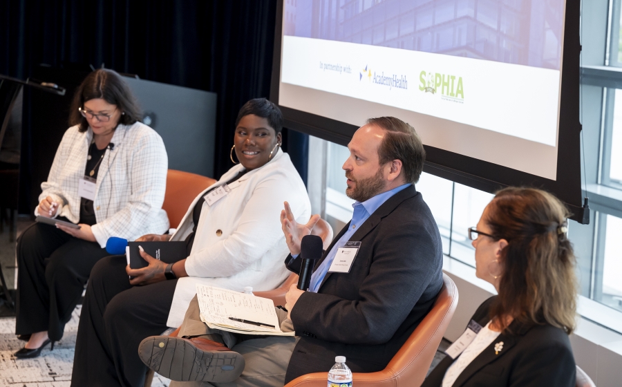Three women and one man are seated in a row with the bottom half of a projector screen visible behind them. The man, Jimmy Dills, is holding a microphone as he speaks.