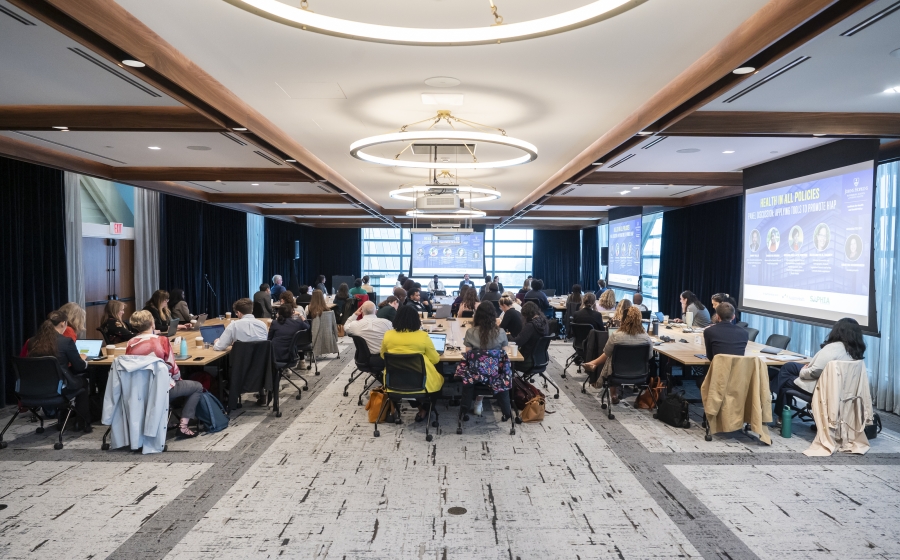 A large room full of people whose backs are facing the camera. They're seated at multiple wooden tables with computers and coffee cups on top. A project screen is against the front of the room and two are against the right wall.