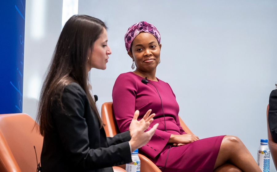 On stage, a woman wearing a purple dress is in focus as she grins at a woman wearing a black blazer.