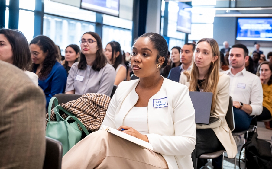An audience member is in focus among a crowd. She's wearing a white blazer and tan pants.