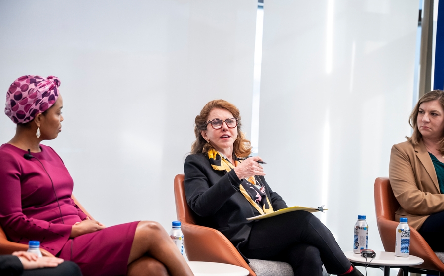 Woman wearing business casual attire is seated on stage between two other women.