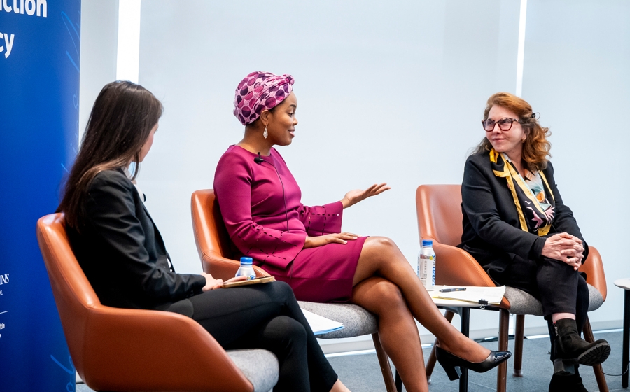 A woman wearing a purple dress speaks with a woman wearing a black blazer on stage.