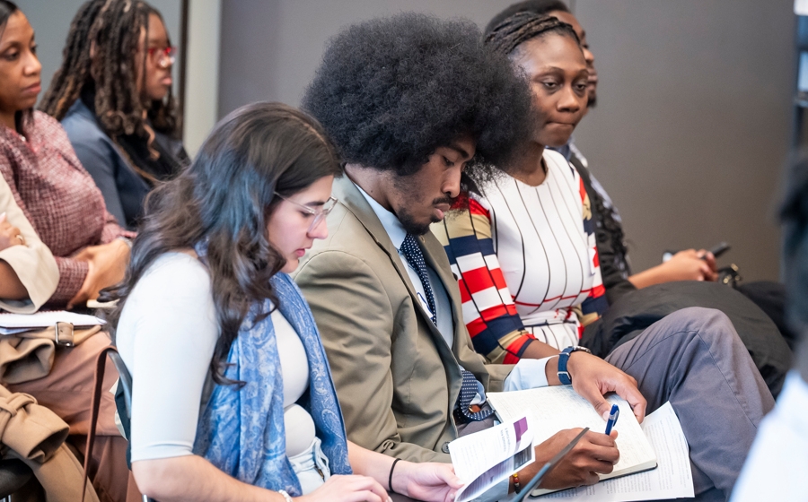 A few audience members are in focus among a crowd. One is typing on her laptop. One is writing in his notebook.