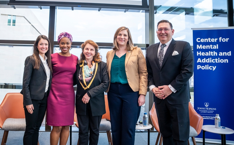 Five adults stand and smile in front of the stage wearing business attire.