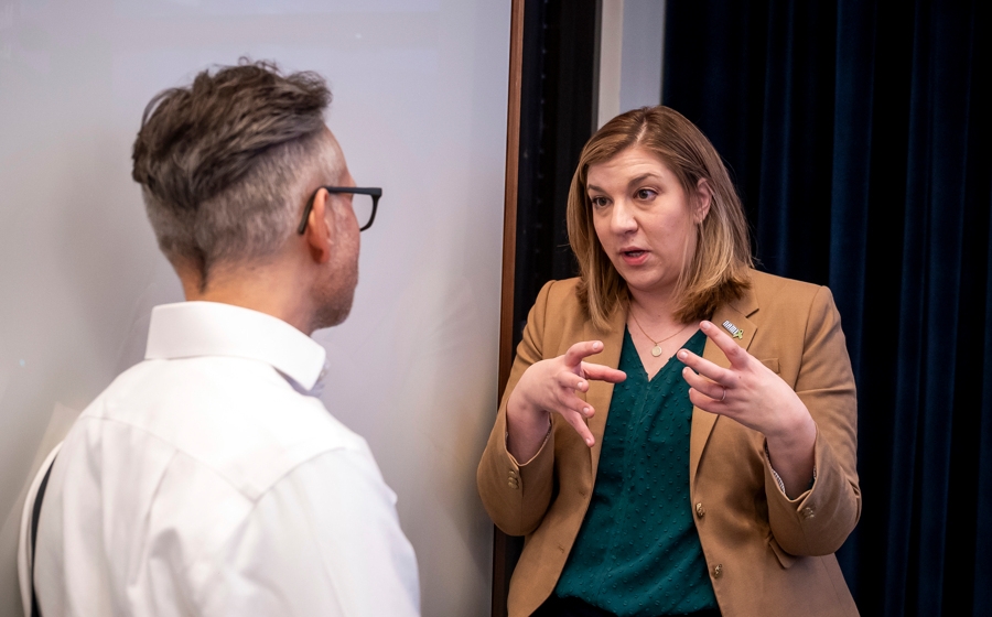A woman wearing a tan blazer speaks to a man. Her hands are mid-air as she gestures.