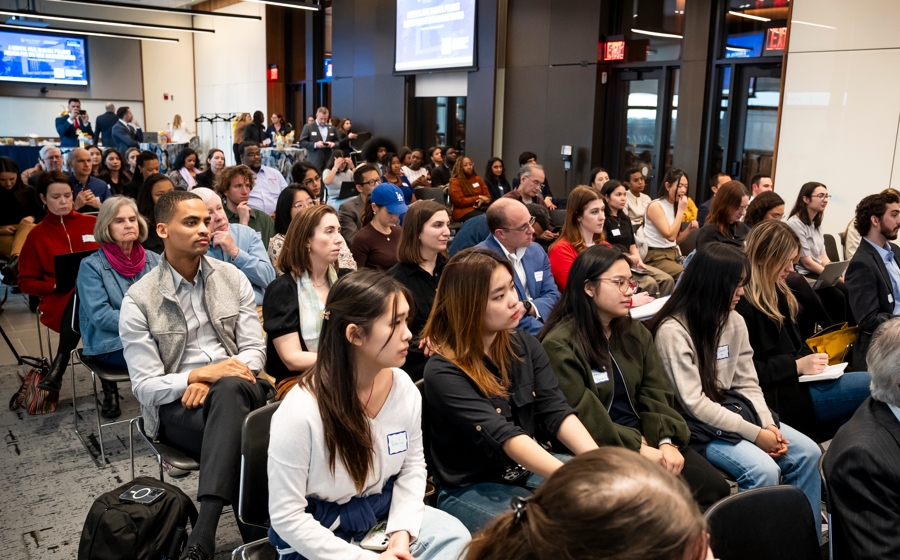 A crowd of over one hundreds people are seated in rows facing the stage.
