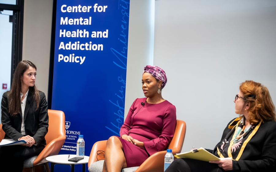 A woman wearing a purple dress and purple headscarf is seated on stage between two other women.
