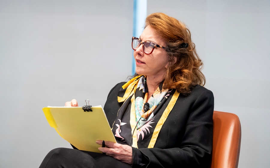 A woman with red hair and a black blazer is seated onstage and holding a yellow folder.