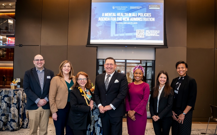 - Seven adults wearing business casual attire stand and smile side by side in event space.