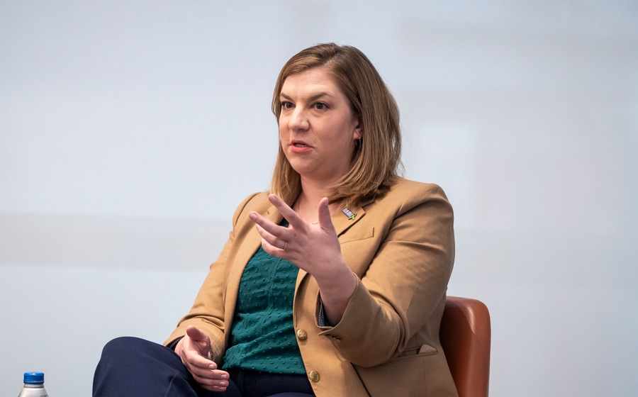 A woman wearing a tan blazer is seated onstage. She's gesturing with her left hand.