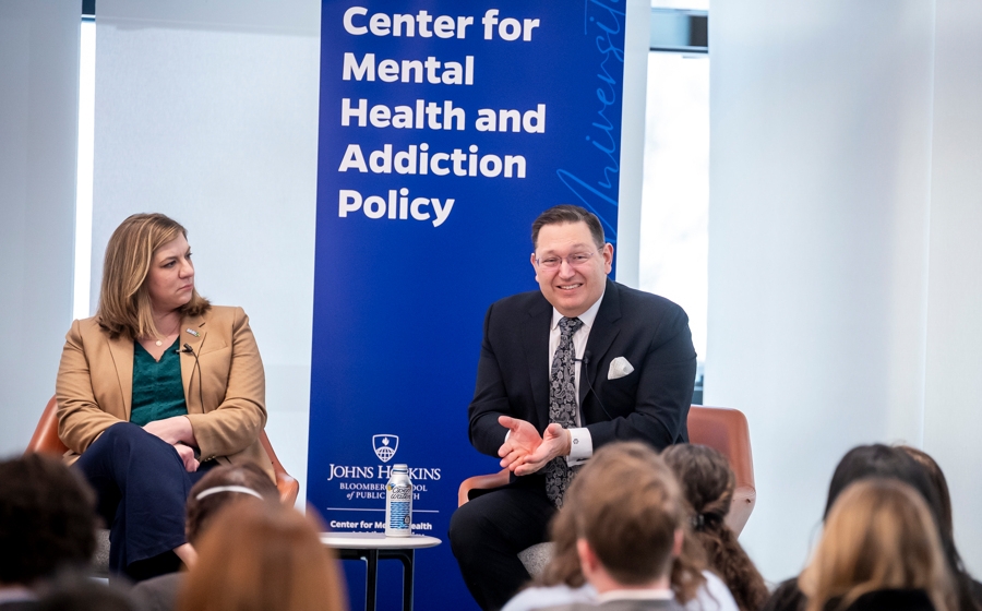 A man wearing a suit is seated onstage. He's smiling and gesturing as he speaks to the crowd.