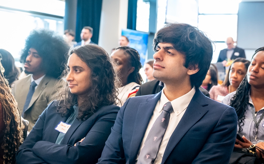 Two audience members are in focus among a crowd. They're wearing business attire.