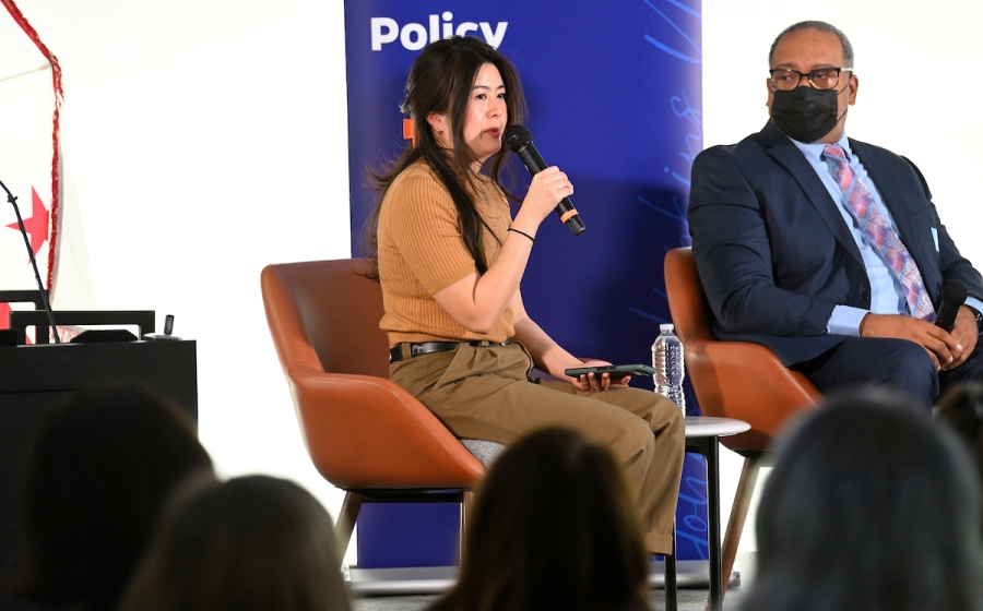 A woman seated on stage speaks into microphone.