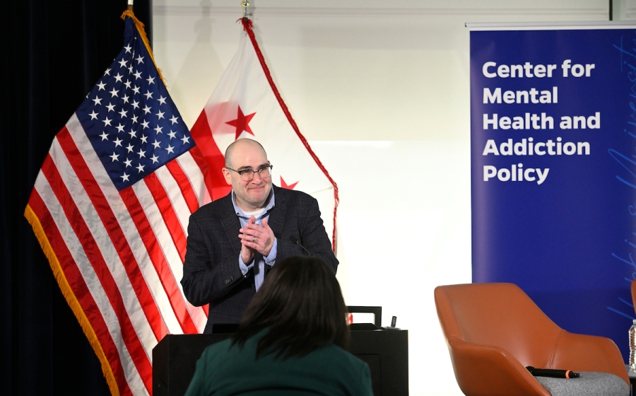 A man smiling and standing behind a podium as he claps.