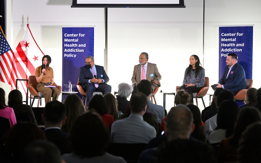 Five adults seated on stage with microphones in hand. One adult seated on far left speaks to audience.