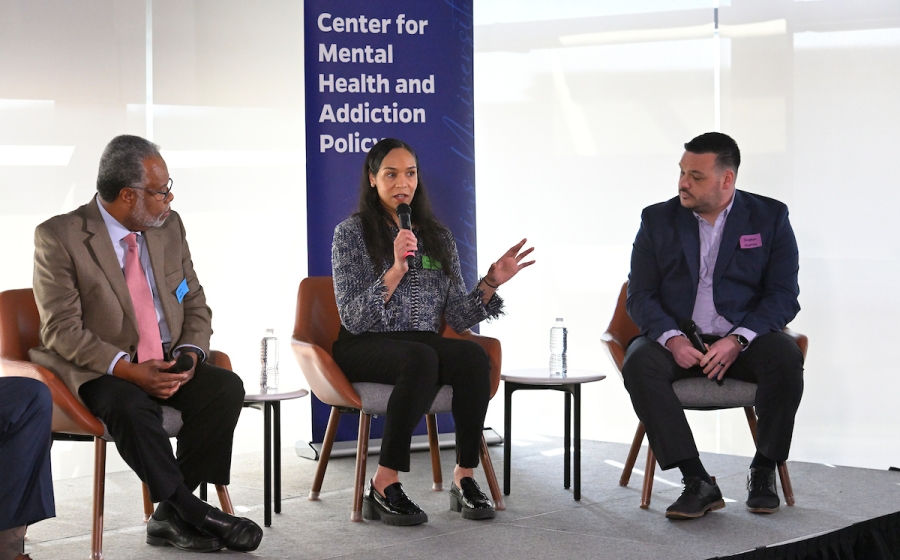 A woman seated on stage speaks into a microphone as she addresses audience.