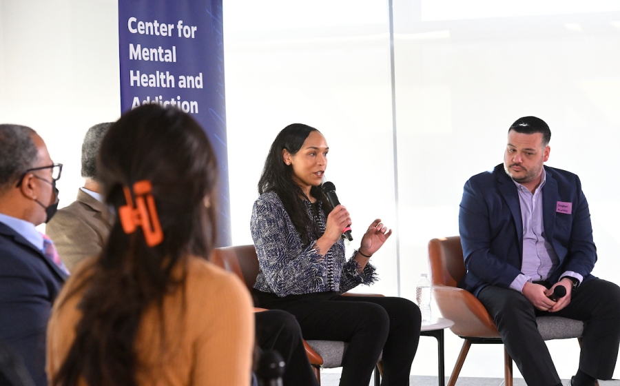 A woman seated on stage speaks into a microphone. Four adults are also seated on stage.