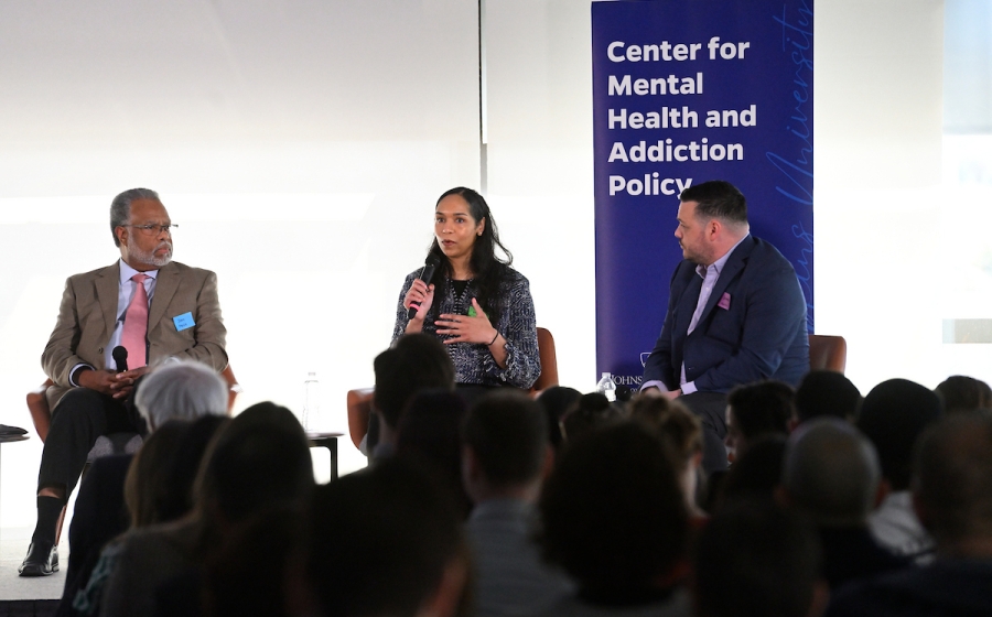 A woman seated on stage speaks into a microphone as she addresses audience.
