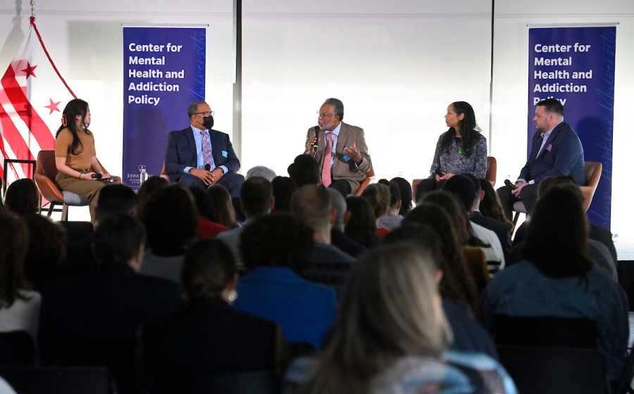 A man seated on stage speaks into a microphone. Two adults are seated to his left and right. Event crowd is visible.