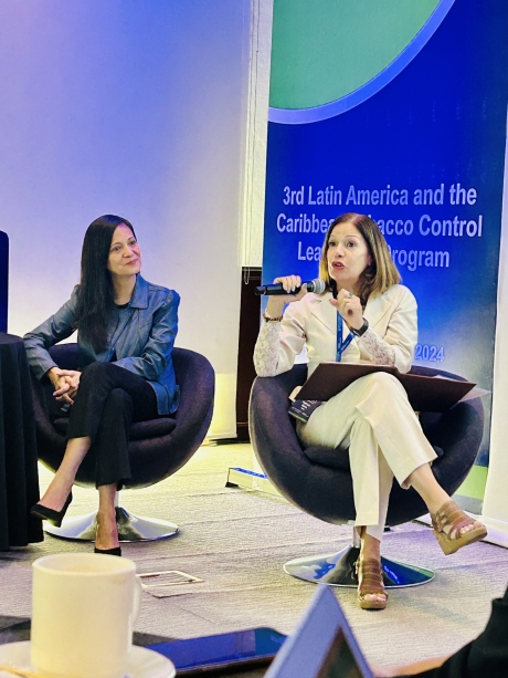 Two women seated on a stage speaking the audience. One wears a white suit. The other wears a blue top and black pants.