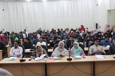 A crowd of people sitting in a large room during a meeting in Bangladesh. Several women with head coverings are in the front row. 