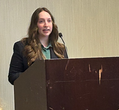 A woman in a dark blazer and green top speaks at a podium