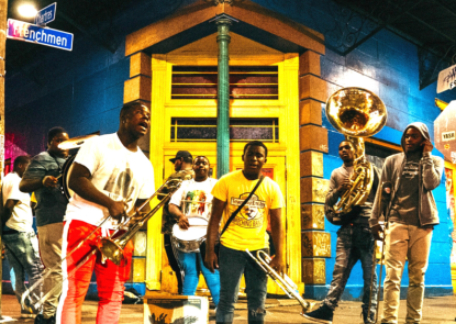 A group of young men holding brass instruments outside a bright yellow and blue building in New Orleans 