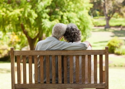 An elderly couple sits on a bench in a park with their backs to the camera.