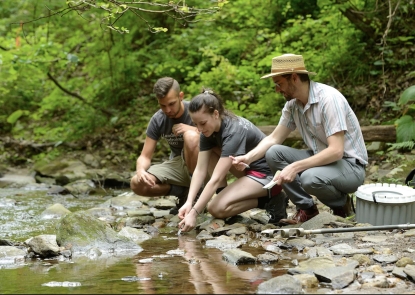 EHE-Harman-students-creek man in hat, young man and young woman take samples from a creek