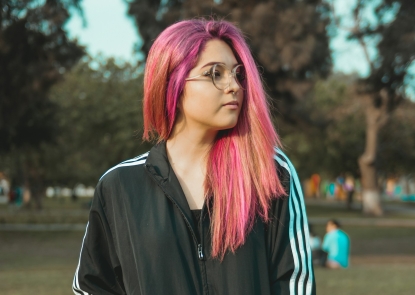 A teenager with long pink hair, standing outside in a park, looking to the distance