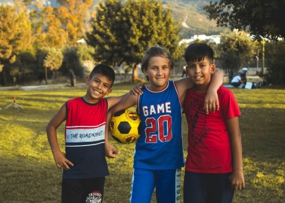 Three boys smiling in a park, one is holding a yellow soccer ball 