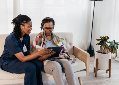 A woman wearing a nurse uniform holds a tablet for an elderly woman to see. They are both sitting on a living room couch.